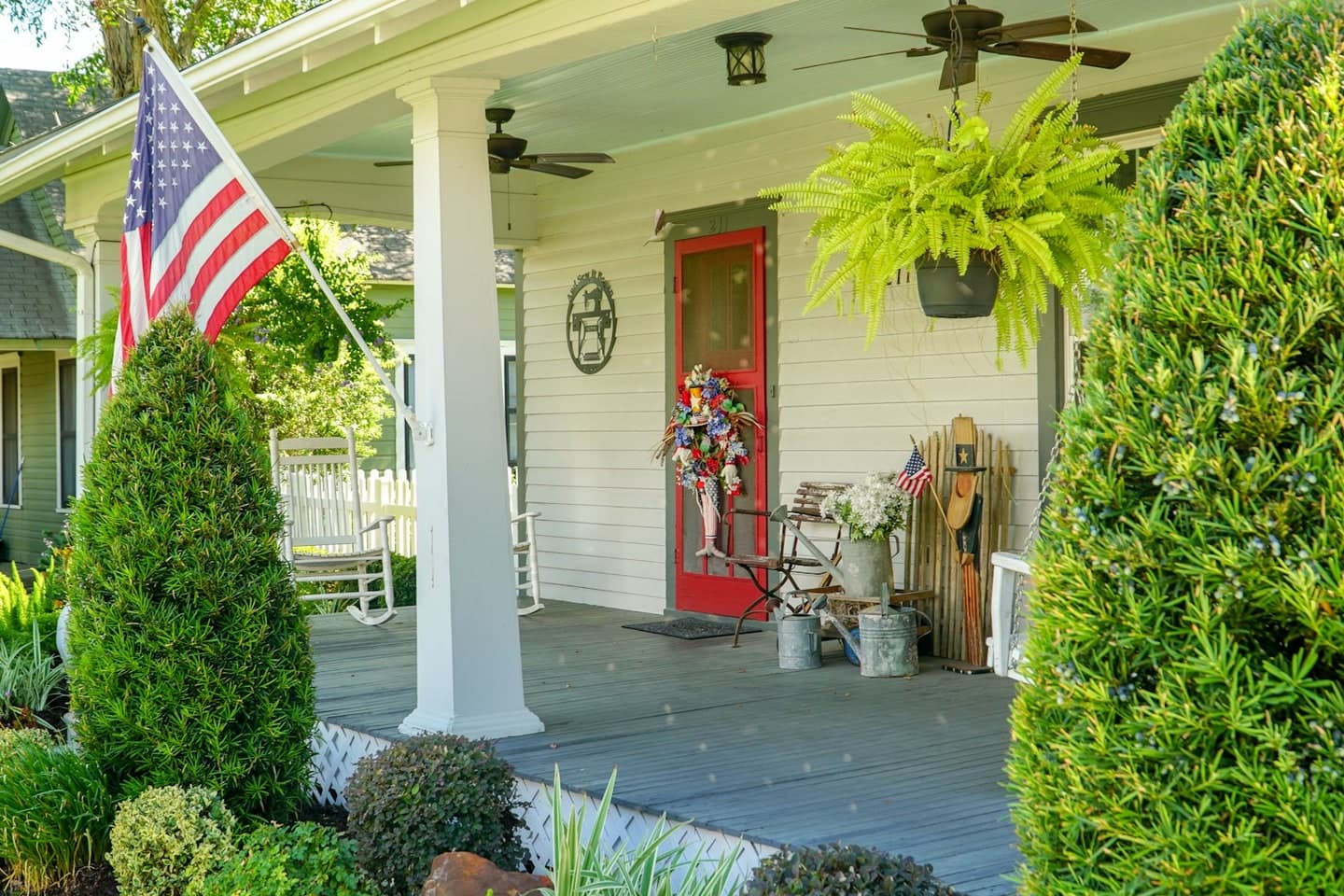 The front porch of a white house with an American flag, hanging ferns and a red door