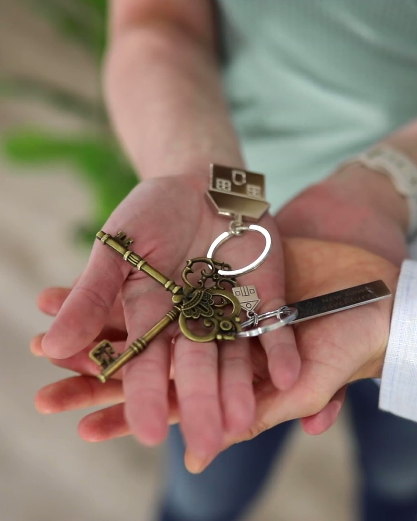 Hands holding house keys and a house-shaped keychain, with a second hand supporting them