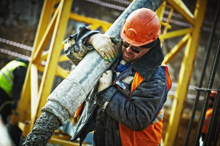 A construction worker in a hard hat carries a concrete hose on a job site