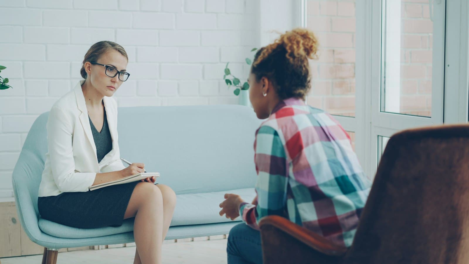 A therapist with a notebook listens to a teenage girl in a bright counseling room