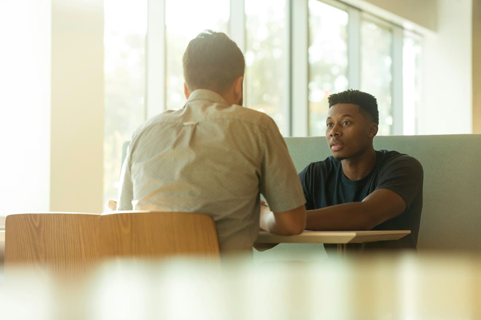 Two men in civilian clothes talk across a table in a sunlit room